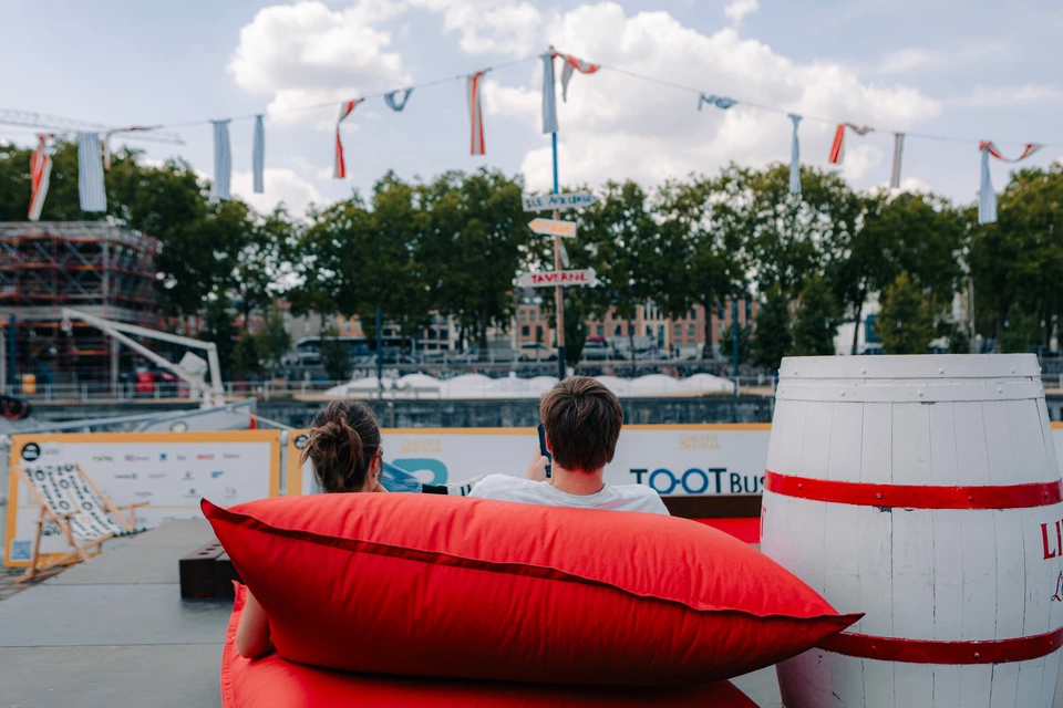 Twee personen ontspannen op rode zitkussens langs het kanaal tijdens Zomerkaai aan de Akenkaai, in een sfeervolle zomeromgeving met zicht op het water en de activiteiten op de kade.